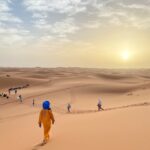 Travelers walking through the vast dunes of Merzouga Desert during a stunning sunset in Morocco.